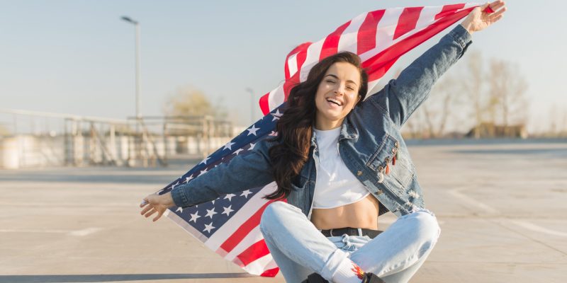 mid-shot-young-woman-holding-big-usa-flag-sitting-leg-crossed